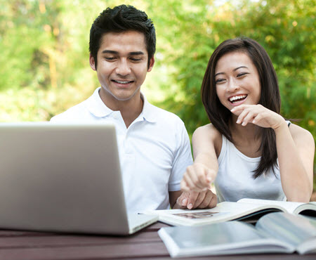 A man and a woman looking at a laptop computer.  both look happy.  The woman has her hand in front of her face.  They appear to be outside as there is bright greenery behing them.  A metaphor for a couple being happy talking about money.