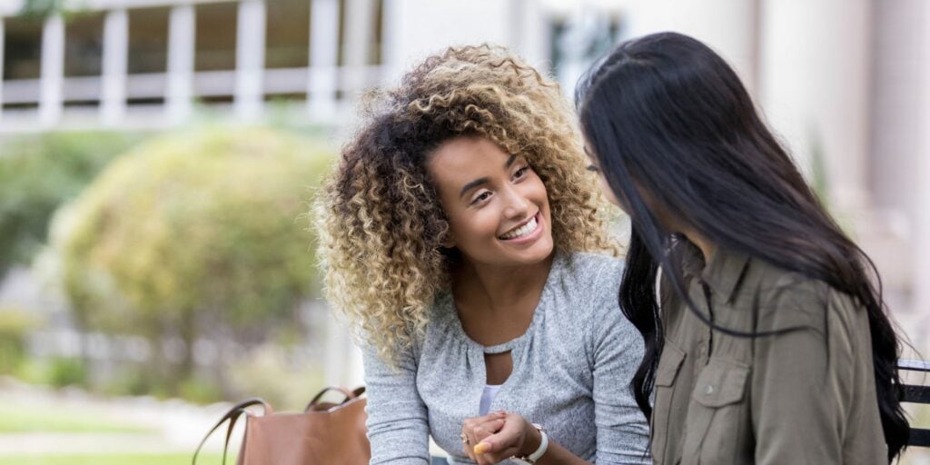 two friends talking on a park bench. The woman facing us is smiling and holding up her hand, we can't see the woman's face who is facing away from us. How to tell your debt or money story from a debt coach.
