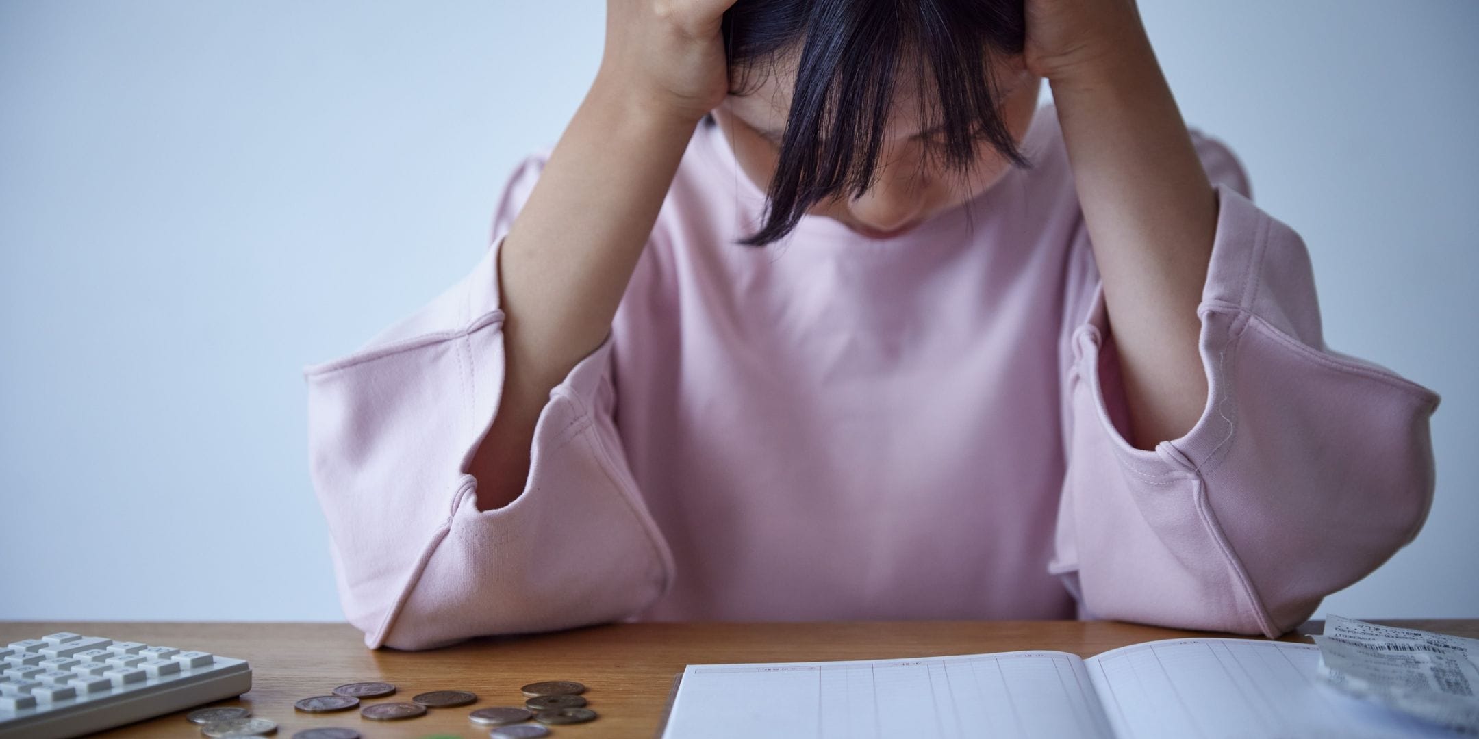 A woman in a pink blouse. She has her head in her hands. On the table in front of her she has some receipts, a spreadsheet workbook, coins and a calculator. Hero image for why budgets don't work