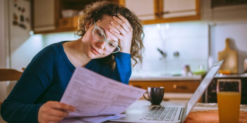 A woman sitting at her kitchen table looking at a bank statement with laptop open in front of her. She looks worried. She can't stop thinking about money.