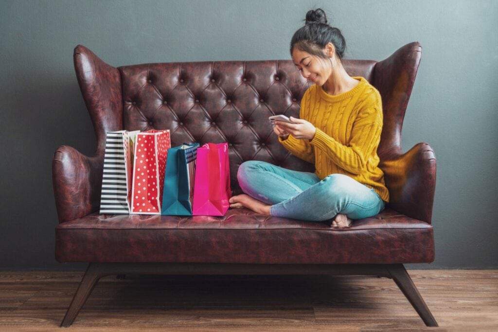 A woman sitting on a small leather couch with four colorful shopping bags next to her. She's sitting cross-legged and looking at her phone. hero image for "why can't I stop spending money"
